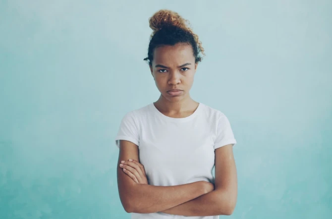 teenage mixed race girl with frizzy hair looking disgruntled at camera in front of light aqua blue background wall